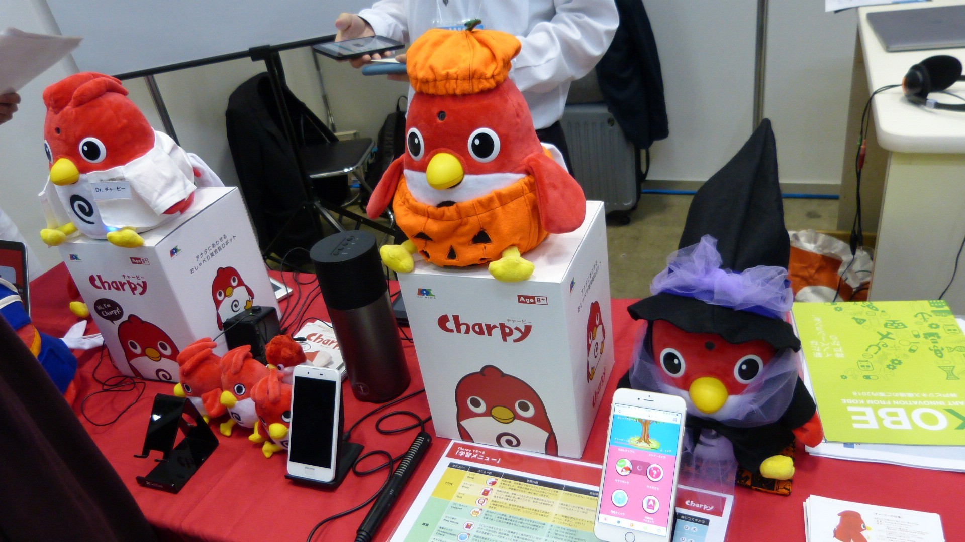 several Charpy toy robot birds with their boxes and some smartphones.  one toy is dressed as a jack-o-lantern and another is dressed as a witch for halloween
